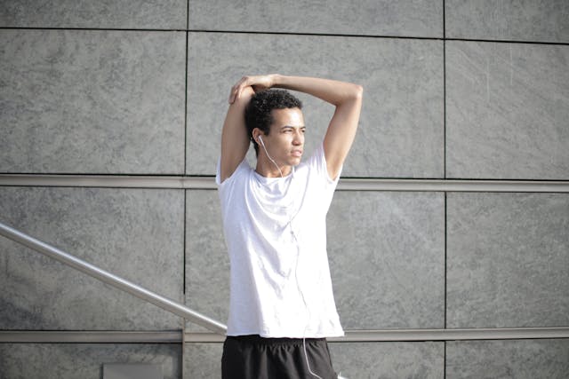 Man stretching arms overhead outdoors while wearing earphones and athletic clothing